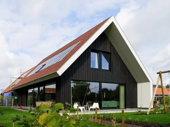 Foto van een landelijke woning met een zwarte houten gevel en rode dakpannen. Er zijn grote raampartijen en daarnaast heeft de woning zonnepanelen.