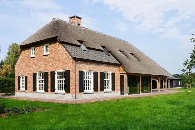 Foto van een moderne boerderijwoning met een rieten kap, klassieke luiken een metselwerk gevel en glazen aanbouw.